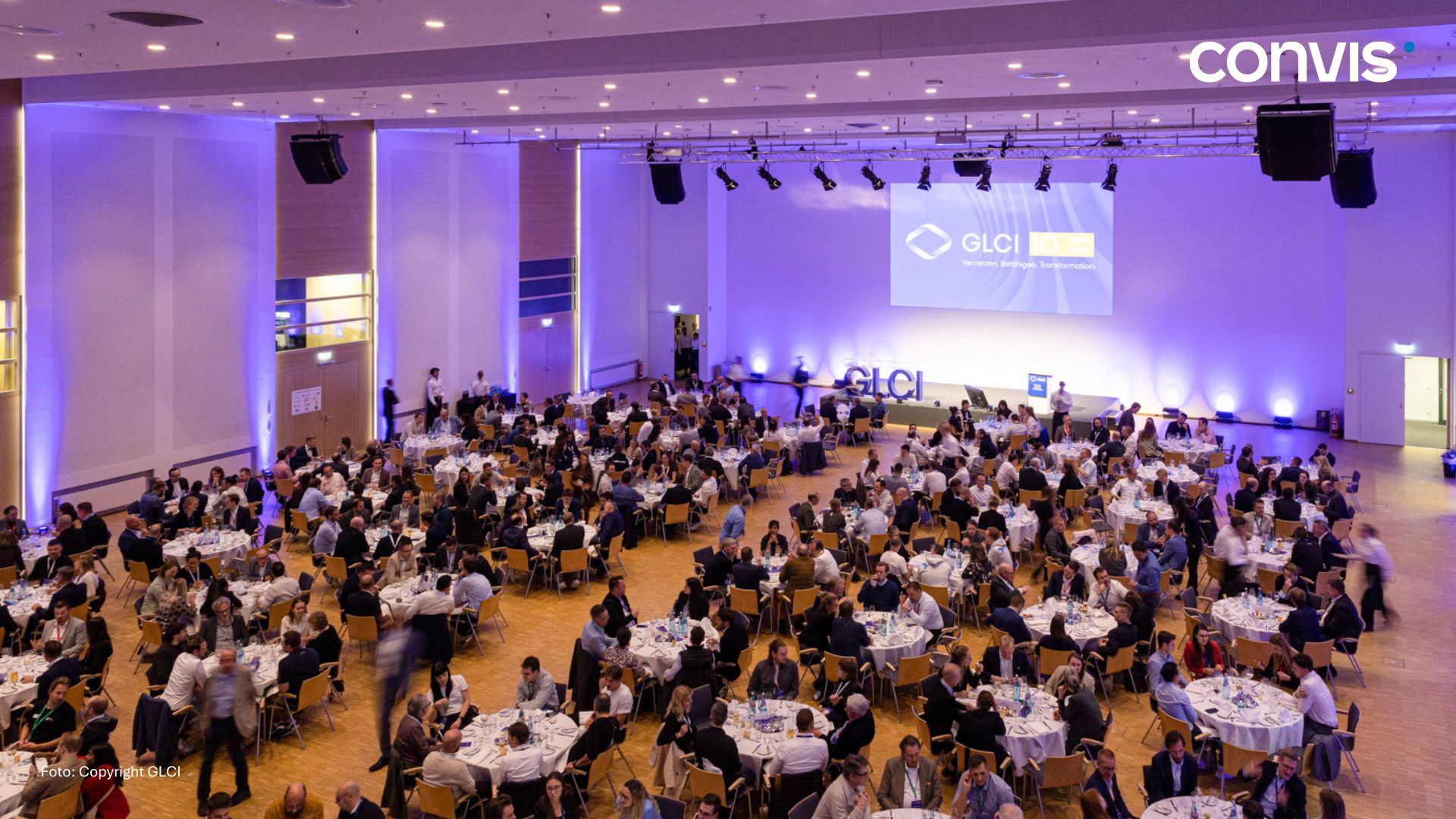 Große Veranstaltung mit festlich gedeckten Tischen und Teilnehmern im Konferenzsaal.