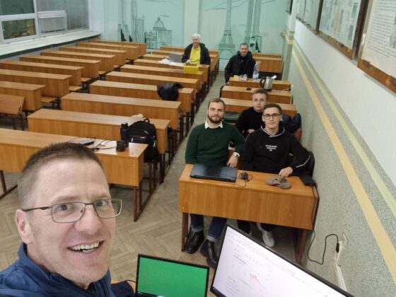 Lehrer und Schüler in einem leeren Klassenzimmer mit Holzschreibtischen und Laptops.