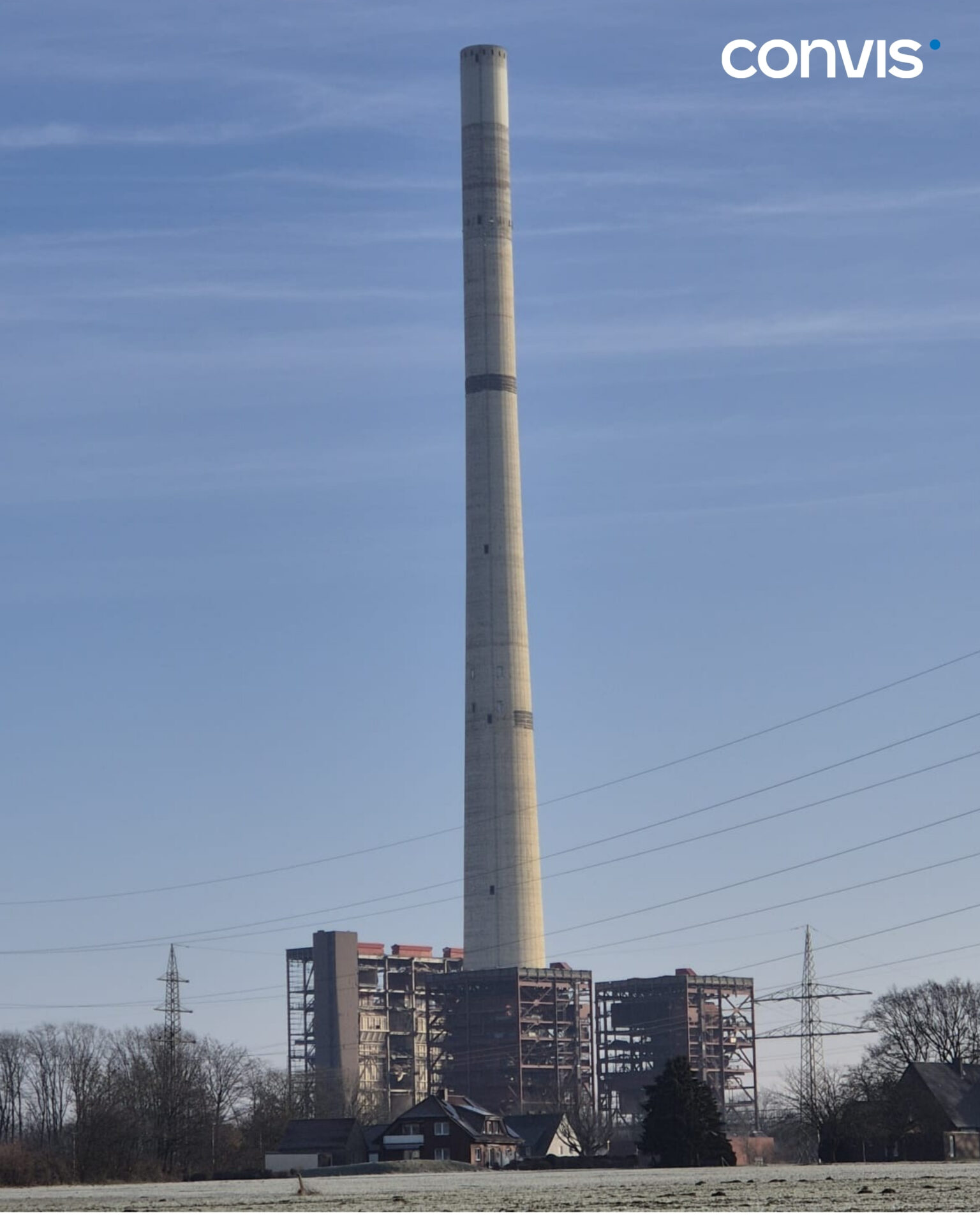 Hoher Schornstein eines Kraftwerks vor blauem Himmel und Stromleitungen im Hintergrund.
