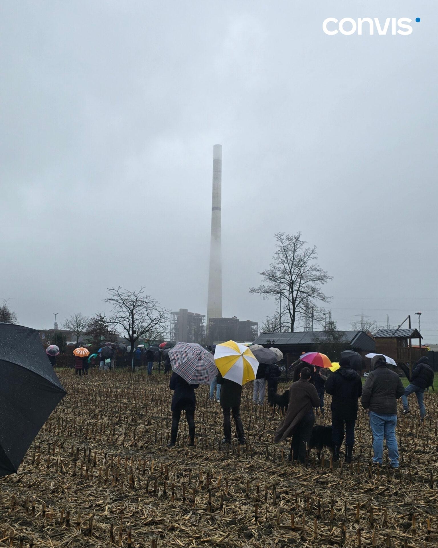 Menschen mit Regenschirmen stehen auf einem Feld vor einem hohen Schornstein.