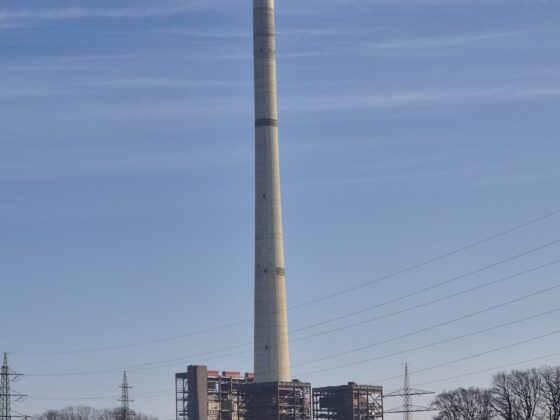 Hoher Schornstein eines Kraftwerks vor blauem Himmel und ländlicher Kulisse.