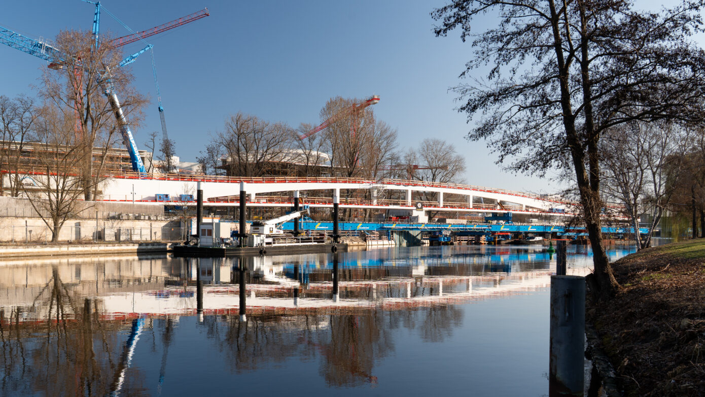 Bauarbeiten am Wasser mit Kränen und reflektierender Oberfläche im Vordergrund.