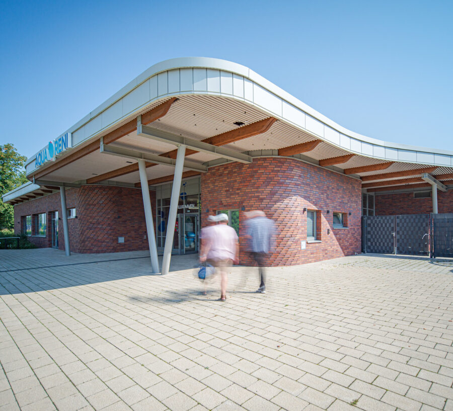 Eingangsgebäude des Kombibad „Aqua Reni" mit geschwungener Fassade, braunen Steinen und Personen im Vordergrund.