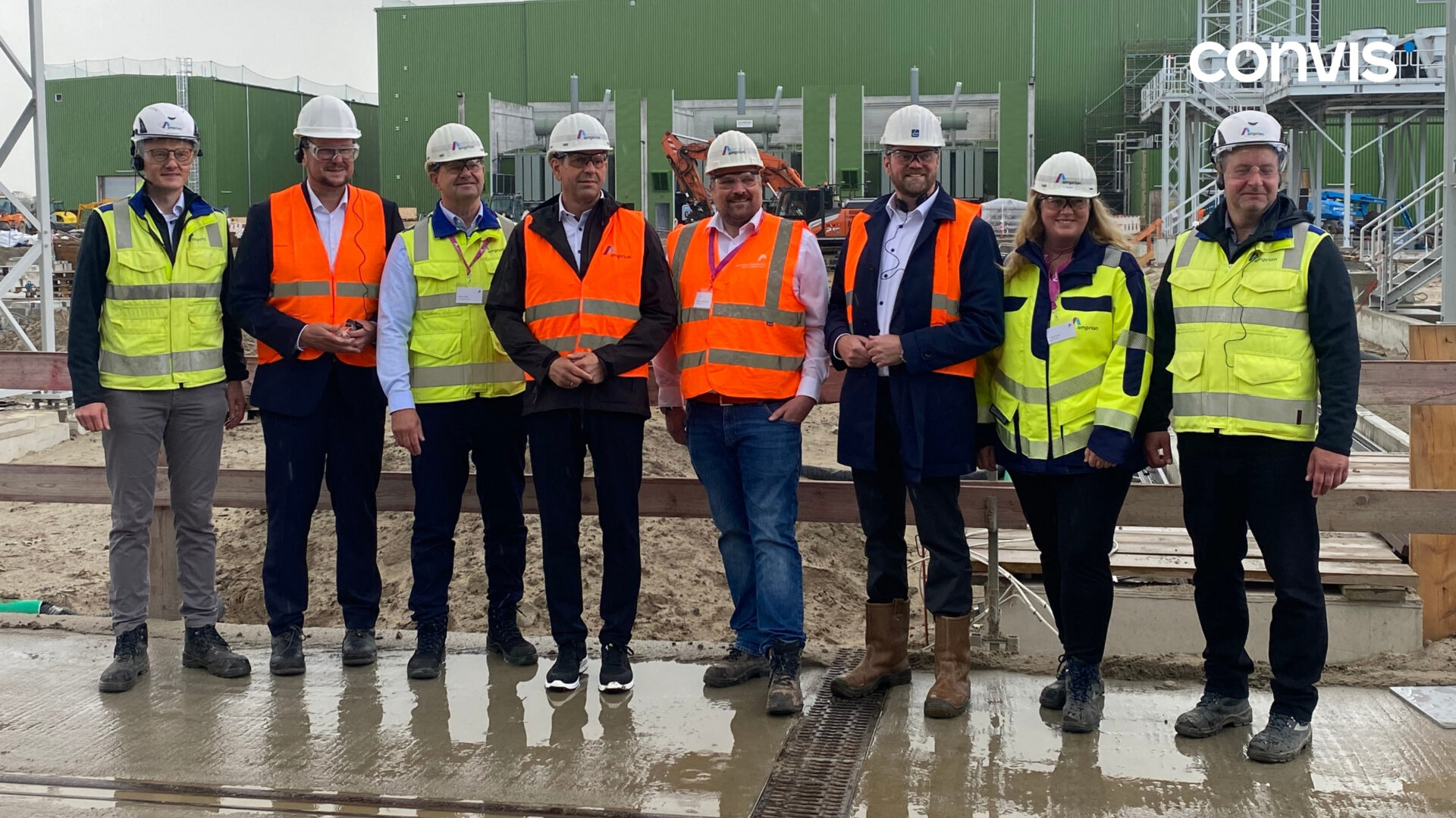 Gruppe von Fachleuten in Sicherheitskleidung auf Baustelle mit grünen Gebäuden im Hintergrund.