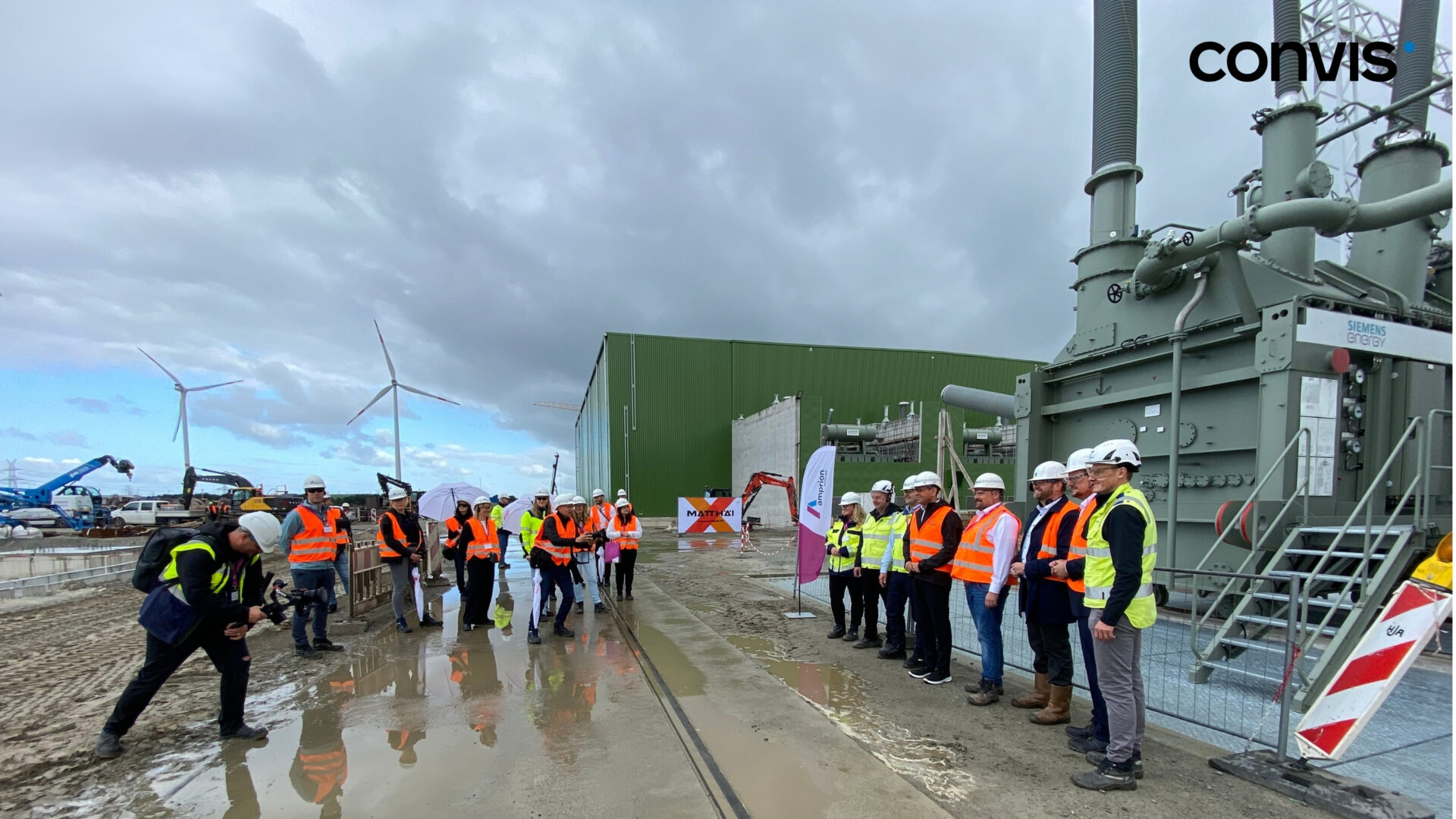 Gruppenteilnehmer in Sicherheitskleidung besichtigen ein Bauprojekt mit Windkraftanlagen im Hintergrund.