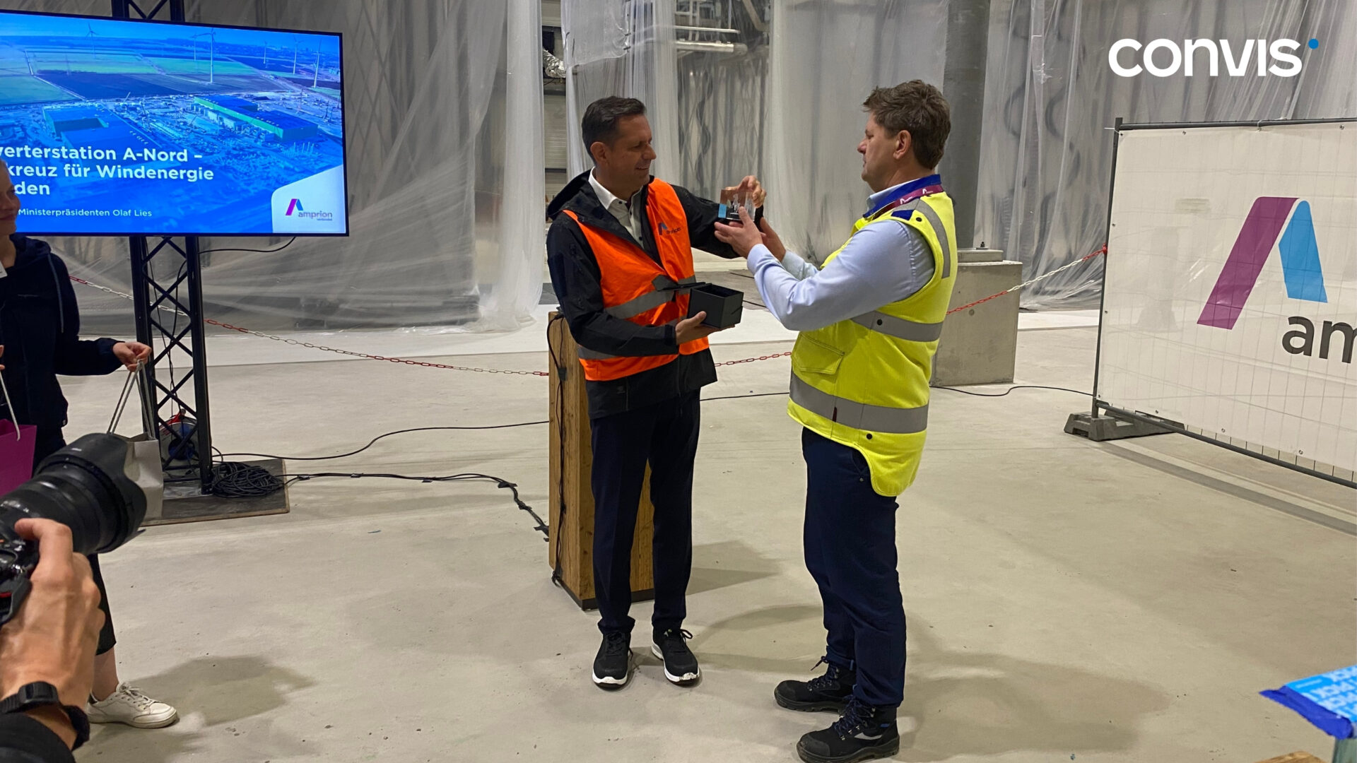 Zwei Männer in Sicherheitswesten präsentieren ein Modell für Windenergie auf einer Baustelle.