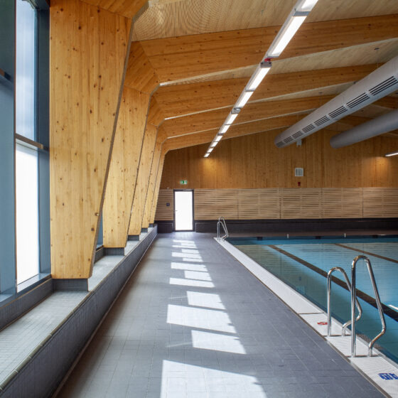 Moderne Schwimmhalle mit Holzdecke und großen Fenstern für Tageslicht.