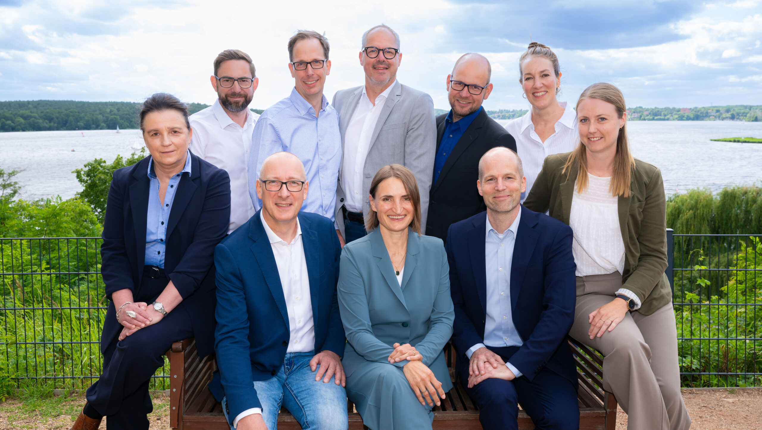 Gruppenfoto von zehn Personen am Wasser, umgeben von Natur und Wolken.