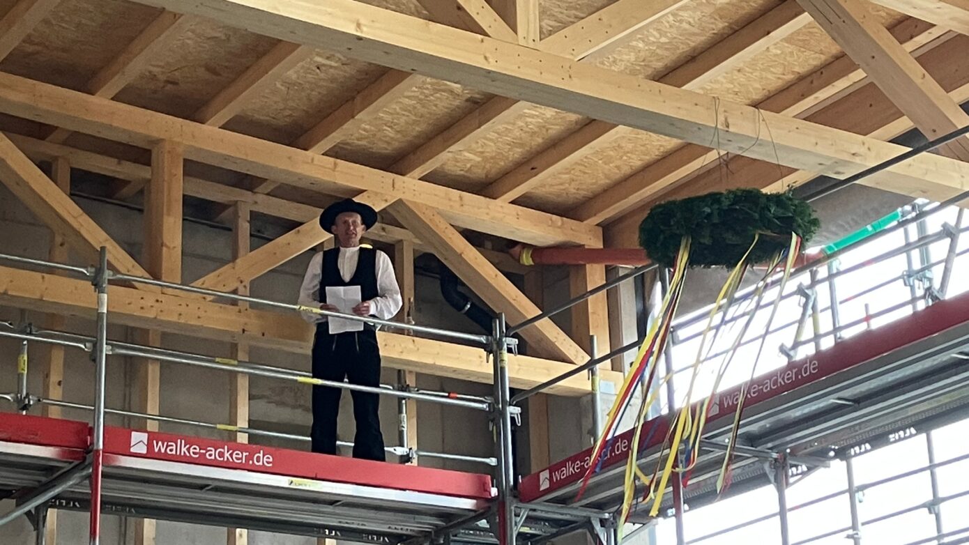 Traditionelle Baufeier: Person in Tracht hält Ansprache auf Gerüst unter Holzdecke.