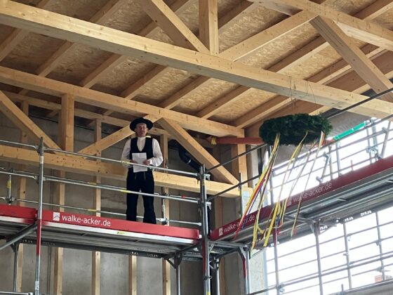 Traditionelle Baufeier: Person in Tracht hält Ansprache auf Gerüst unter Holzdecke.