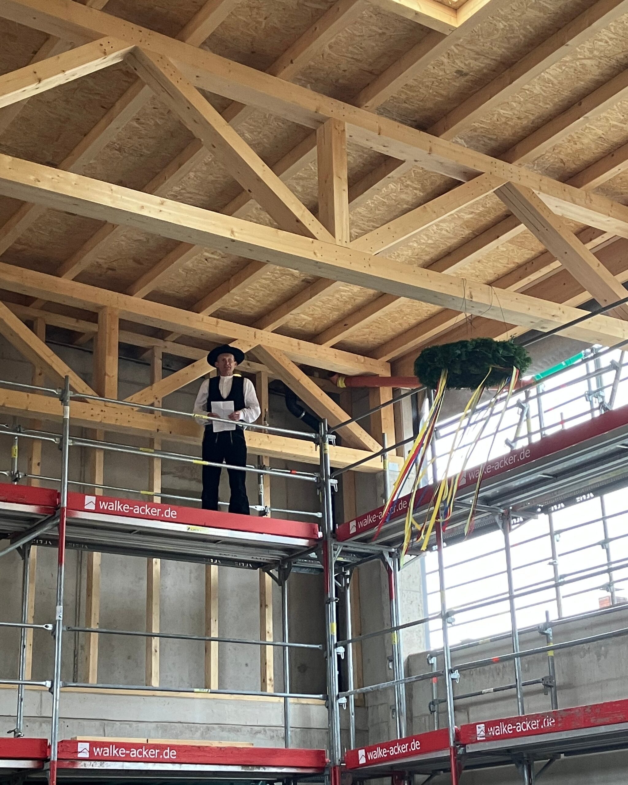 Traditionelle Baufeier: Person in Tracht hält Ansprache auf Gerüst unter Holzdecke.