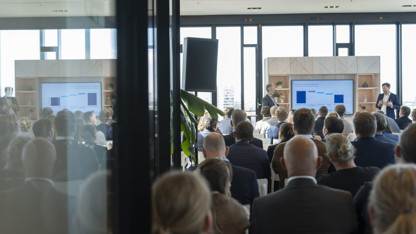 Konferenz mit Präsentation vor Publikum in modernem, lichtdurchflutetem Raum.