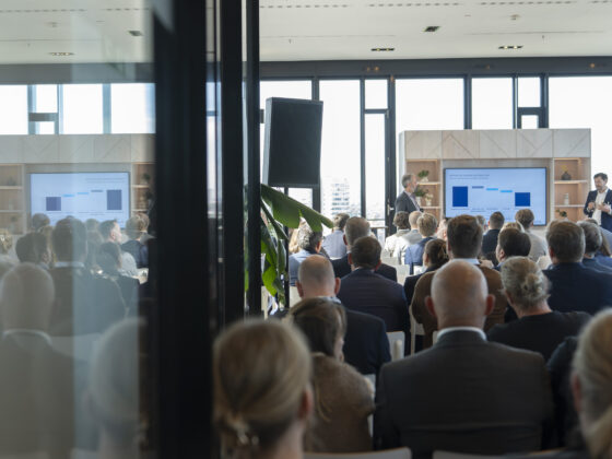 Konferenz mit Präsentation vor Publikum in modernem, lichtdurchflutetem Raum.