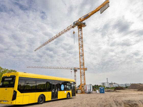 Gelber Bus vor Baustelle mit Kränen und bewölktem Himmel.