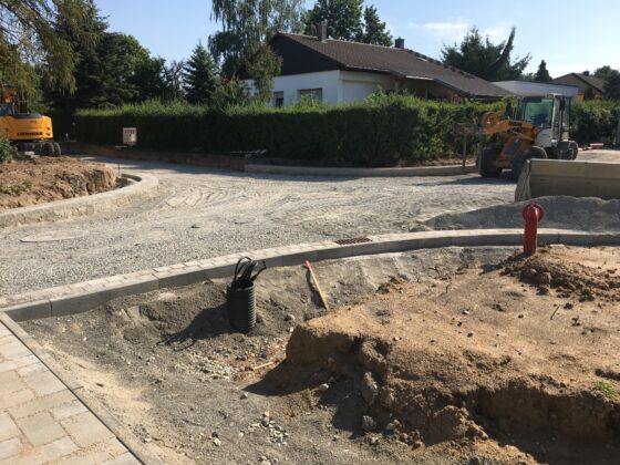 Bauarbeiten an einer Straße mit Maschinen und unbefestigtem Untergrund in Wohngebiet.