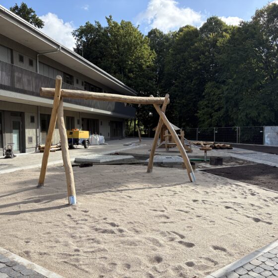 Schaukel auf einem Spielplatz in der Bauphase, umgeben von Bäumen und Sand.